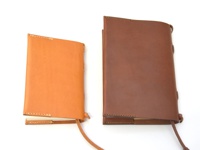 兩個職人手作皮革附繩書套，左邊為淺棕色，右邊為深棕色。兩款書套皆為皮革材質，邊緣有縫線處理，並附有可綁繫的皮革繩。