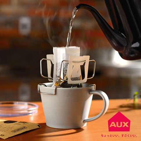 銀色金屬可調式耳掛咖啡架，固定著兩個濾掛式咖啡包，架子夾在一個淺灰色陶瓷馬克杯的邊緣。熱水從黑色細嘴水壺中倒入咖啡包，產生蒸汽。