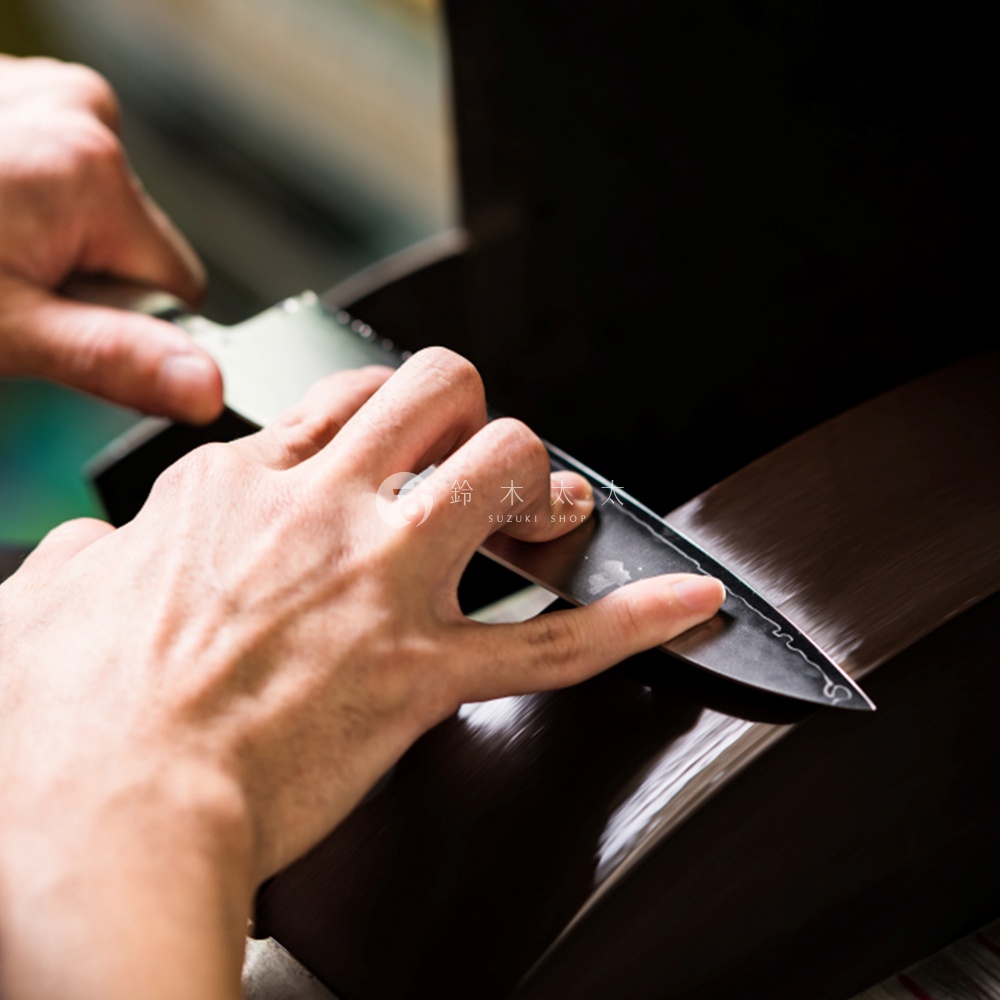 手持日本刀，在拋光工具上打磨刀刃，刀身上印有「鈴木太」和「SUZUKI SHOP」的標誌。