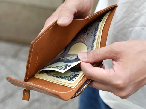 一雙手正在操作一個棕色皮革方形短夾，夾內有日圓紙鈔。短夾具有拉鍊和皮革拉片，縫線清晰可見。