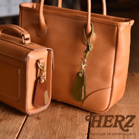 兩款TAN色系皮革包，其中一款為手提包，另一款為小方包，皆有附上金屬扣環及皮革吊飾，吊飾由棕色與綠色皮革及金屬片組成。