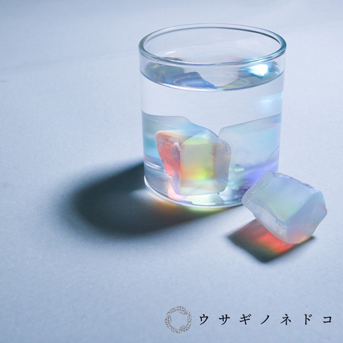 玻璃杯中盛有透明液體，杯內及杯外各有一塊不規則的稜鏡石，呈現虹彩色澤，主要為粉紅、黃、藍、紫等光影變化。