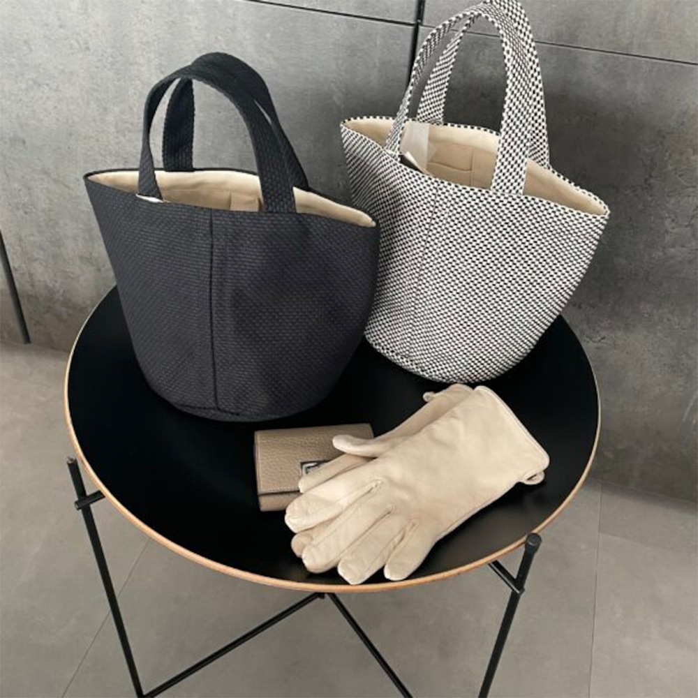 兩款三河木綿刺子織圓底水桶包，一款為黑色，另一款為黑白格紋。包包內襯為米白色。桌上另有米色皮革短夾和一雙米色皮革手套。