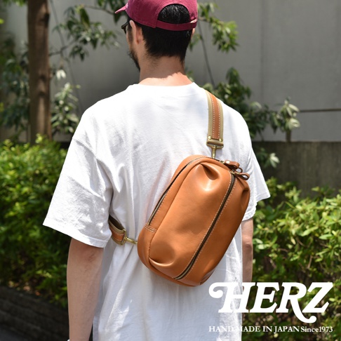 日本HERZ職人手作皮革立體運動斜背包，棕色牛皮革材質，斜背於男性身上，品牌HERZ和HAND MADE IN JAPAN Since1973字樣於右下角。
