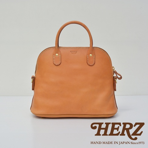 經典貝殼造型手提包，採用棕褐色皮革製作，配有雙提把與側邊拉鍊，包身正面有品牌壓印標誌。