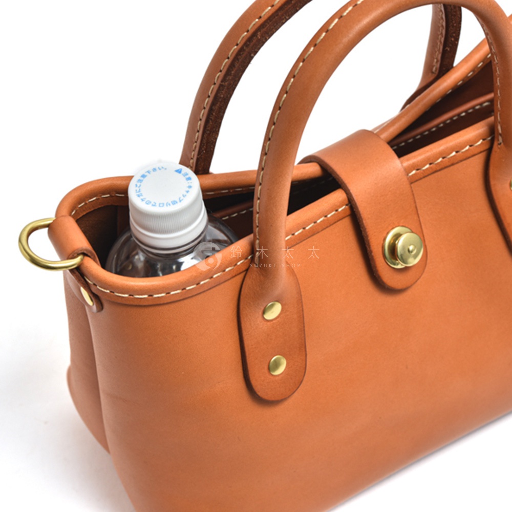 職人手作皮革迷你托特包，採用棕褐色光滑皮革製成，配有圓形提把、金色釦環和金屬旋釦。包內放置一瓶礦泉水，突出包款的尺寸與實用性。
