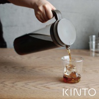 手持深棕色把手的透明玻璃冷泡壺，正將深色液體倒入裝有冰塊的玻璃杯中。