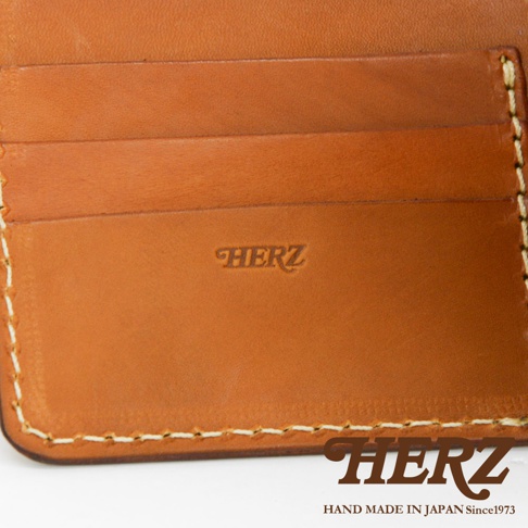 職人手作皮革二折橫式卡片紙鈔小短夾，近距離特寫，展現溫潤的棕色皮革材質、多個卡片夾層，以及壓印的「HERZ」品牌標誌和縫線細節。