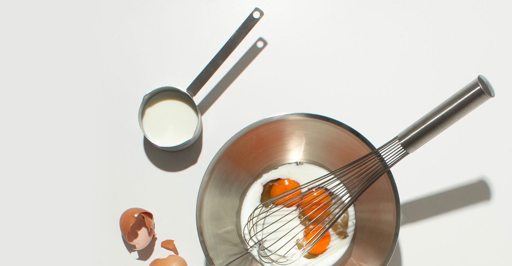 不鏽鋼雙口長柄計量杯，內含牛奶，旁邊是不鏽鋼打蛋盆，盆內有雞蛋和牛奶，以及蛋殼碎片。