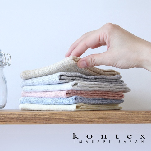 一疊 MOKU LINEN 輕薄速乾棉麻拭巾，由上至下顏色包含淺黃、淺灰、白色、淺粉、淺藍、深灰、卡其色。一隻手正在拿起最上面一條淺黃色拭巾。