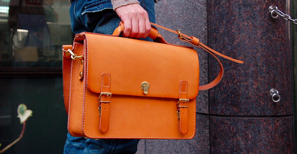 側背著一個橘棕色皮革製成的公事包，包款有提把和可拆卸的斜背帶，正面有三個金屬扣環和一個旋轉鎖。