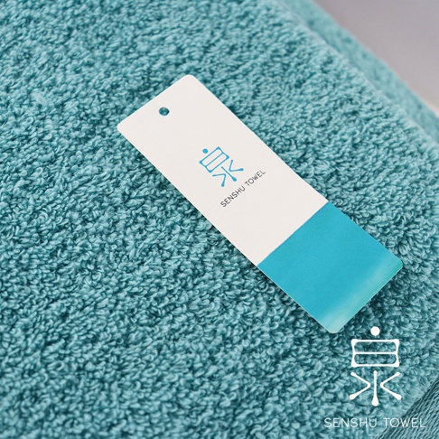 一條藍綠色長纖迷你浴巾，材質柔軟，表面有細緻的毛圈，上面放置著一張白色吊牌，印有藍色品牌標誌及「SENSHU TOWEL」字樣。