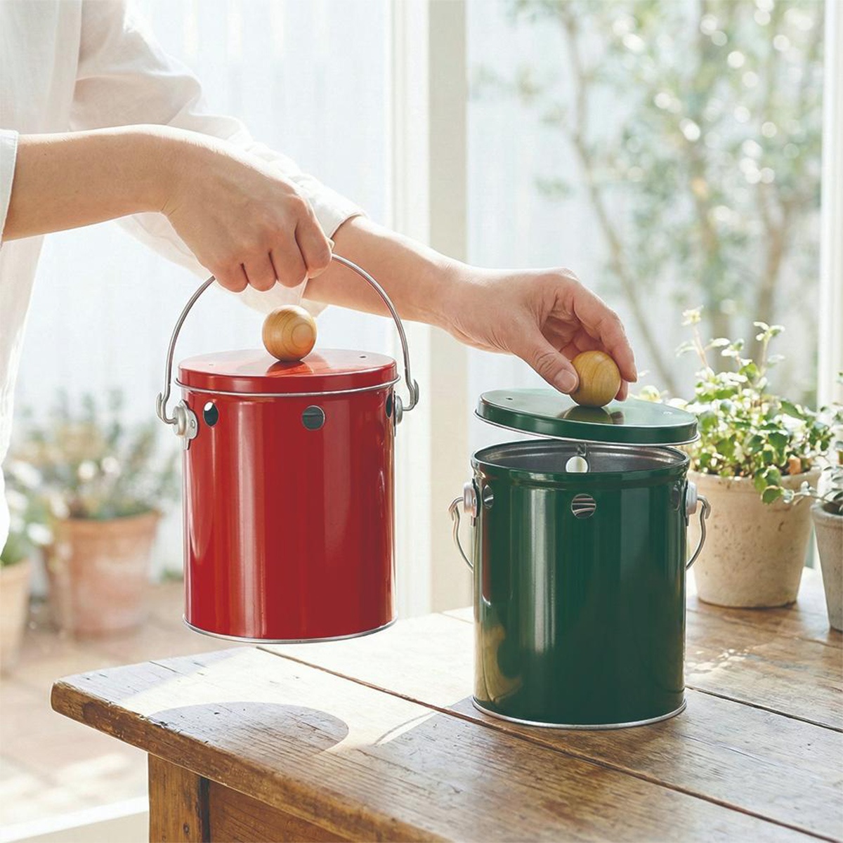 兩個人手持紅色和綠色的 OBAKETSU 手工製蚊香桶，桶身由鍍鋅鋼製成，帶有木質提手和蓋子。