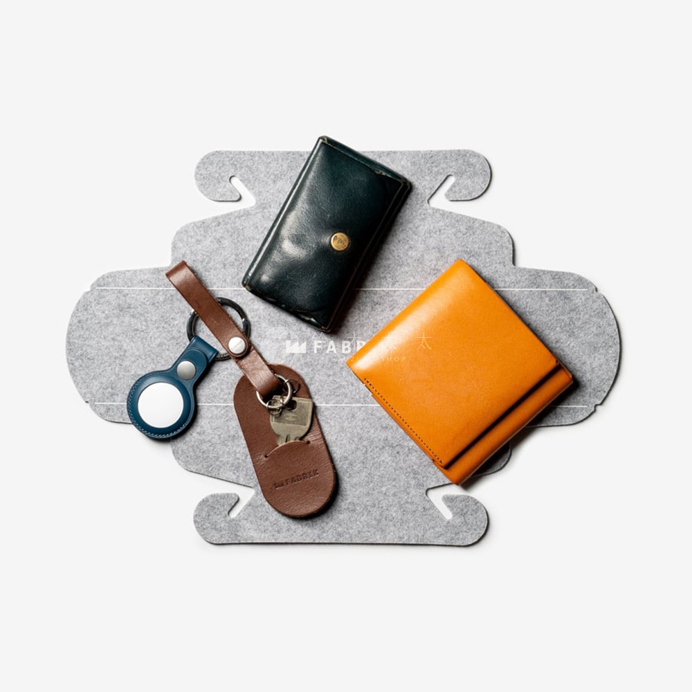 FABRIK 皮革隨身小物，包含一個深綠色長夾、一個橘色皮夾、一個附有鑰匙的皮革鑰匙包，以及一個藍色 AirTag 保護套，皆擺放在灰色毛氈收納墊上，由上往下拍攝。