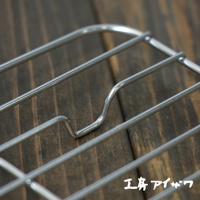方形不鏽鋼瀝油架，由多條銀色金屬線條編織而成，呈現簡潔的網格狀結構，部分線條彎曲成U形，架設於深棕色木紋背景上。