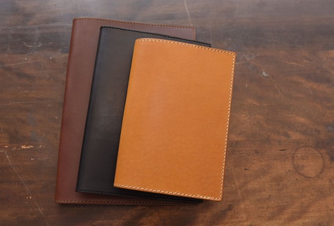 三本職人手作皮革A5筆記本書套，分別為深棕色、黑色和淺棕色，皮革材質，車縫線精緻，擺放在木質桌面。