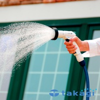 手持白色灑水槍，噴出細緻水霧，槍身有「NOZZLE 5」字樣，連接藍色軟管，背景為綠色窗格和磚牆。