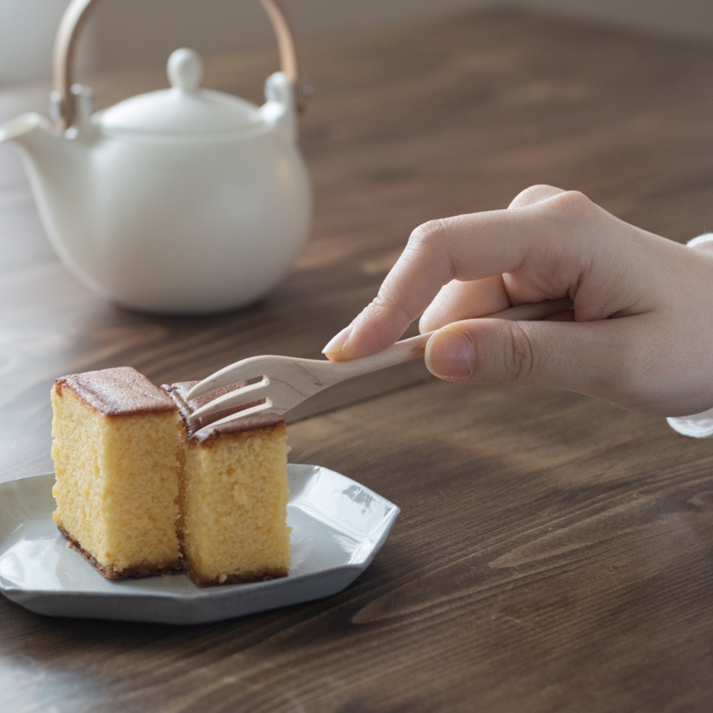 一隻手拿著楓木點心叉，叉著兩塊金黃色的蛋糕，蛋糕放在白色六角形盤子上，背景是白色的茶壺和木質桌面。