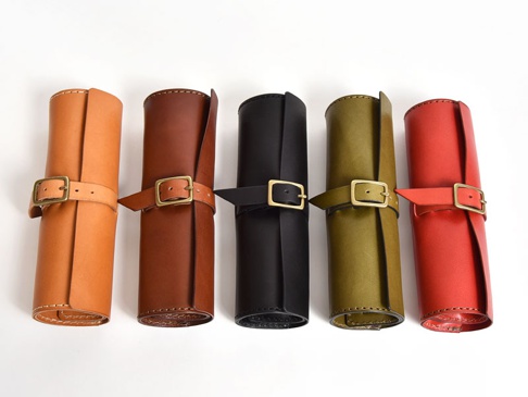 五款職人手作皮革捲捲收納筆袋，顏色分別為淺棕、深棕、黑、墨綠與紅色。筆袋均為圓筒狀，以皮革包裹並以皮帶及銅釦固定。