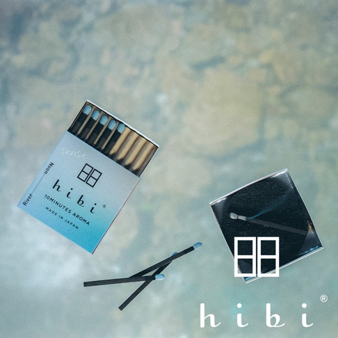 hibi香氛火柴禮盒平放在淺藍色水面上方，左側為印有品牌名稱與10分鐘香氛字樣的藍色包裝盒，右側為放置已點燃冒煙火柴的黑色底座，畫面下方有兩支火柴交叉擺放，整體呈現簡約清新的日式設計風格。