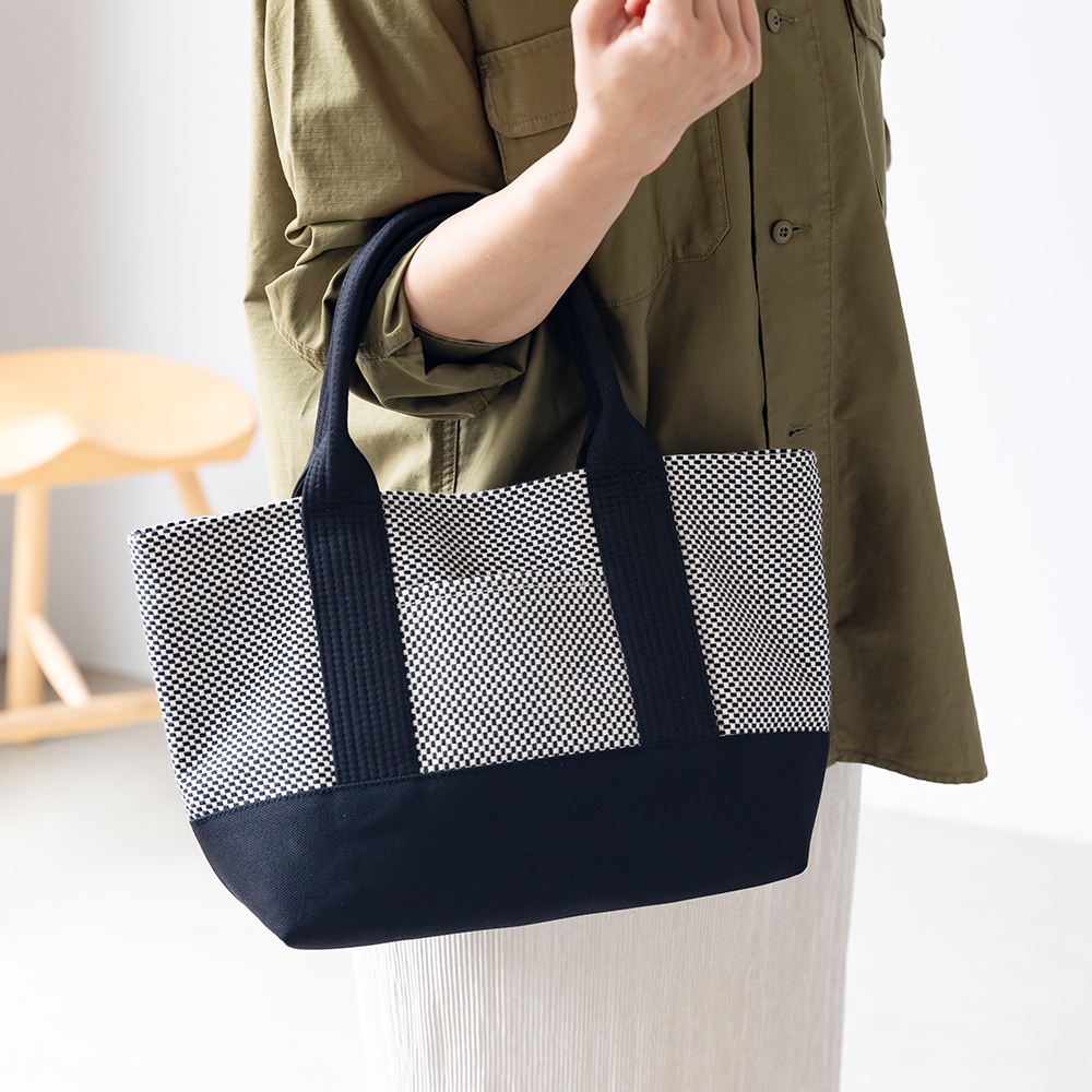 一位女士肩背一個三河木綿刺子織迷你托特包。包包上半部為黑白棋盤格圖案，下半部為純黑設計，配有黑色織帶提把。