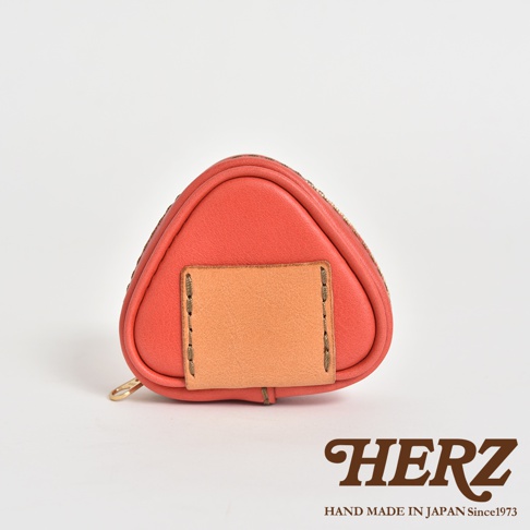 橘紅色皮革製成的三角飯糰造型小物收納包，正面有一個淺棕色皮革裝飾片，以縫線固定。