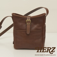 職人手作的棕色皮革輕便單肩側背包，包身方形，配有可調節的肩帶和一個金色帶扣的翻蓋設計，前方有一個方形貼袋，整體風格簡約。