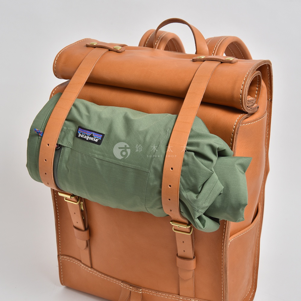 職人手作皮革露營登山用大容量後背包 R-19，採用鞣製的棕色皮革製成，配有可調節的皮革揹帶和捲蓋式頂部，外部固定了一個綠色的收納包，上面印有 Patagonia 品牌標誌。