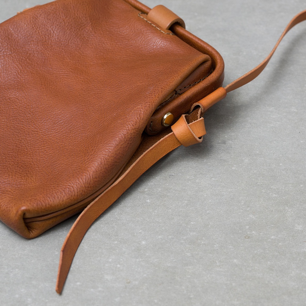 職人手作皮革Organ迷你杜勒斯側背包，採用柔軟的棕褐色皮革，設計簡約，附有細長的皮革背帶，以皮革繩結固定。