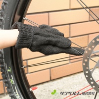 一隻戴著黑色針織清潔手套的手，正觸碰自行車輪的鋼絲。手套表面有細小的網格紋理。背景是淺棕色的磚牆。