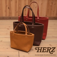 三款職人手作皮革托特包，由左至右分別為駝棕色、深棕色與紅色。三款包皆為簡約設計，採用光滑皮革材質，配有相同色系的皮革提把，包身點綴著金屬卯釘。