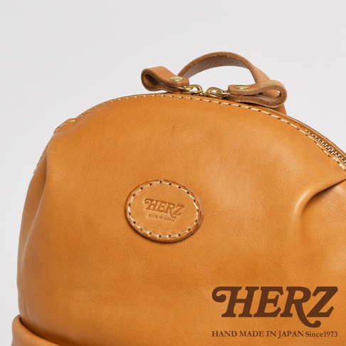 近拍特寫，展示一個 tan 色全皮革材質的圓角後背包，包包上有「HERZ」的壓印標誌，金屬拉鍊細節，以及「HERZ HAND MADE IN JAPAN Since1973」的文字品牌標示。