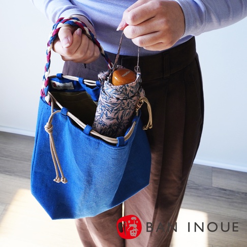BAN INOUE 麻花辮束口手提包，採用藍色帆布製作，搭配印有綠葉圖案的摺疊傘，手提包的提把為編織設計，側面有抽繩束口細節。