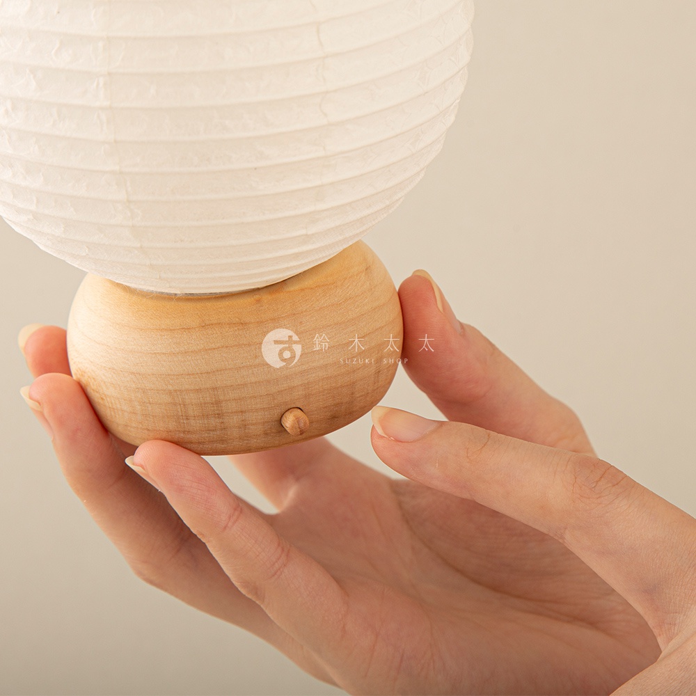 一隻手捧著一個迷你裝飾提燈，提燈上半部是白色紙質燈罩，下半部是圓形淺木色木質底座，上面印有日文和英文的品牌標誌，底座側面有一個小小的木質開關。