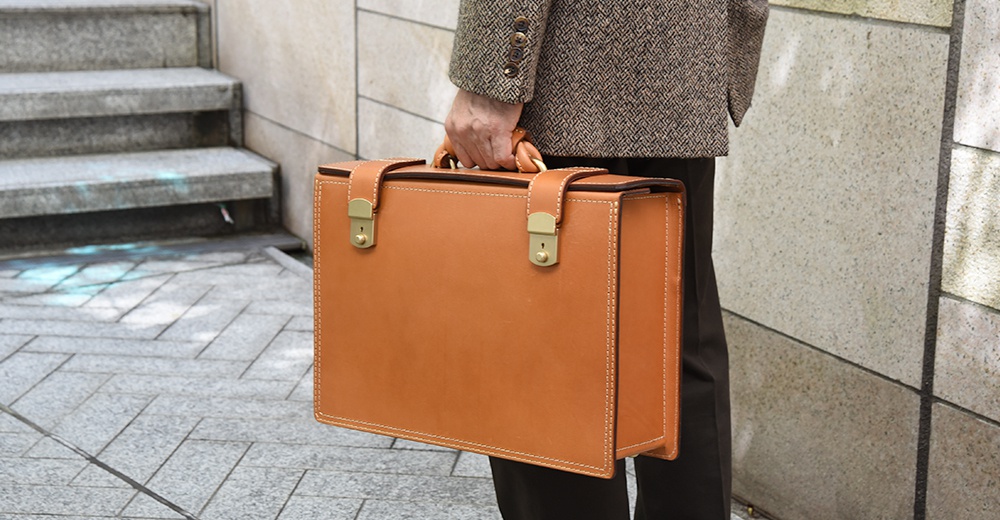 職人手作的咖啡色皮革機師箱，箱體方正，設有兩組金色金屬鎖扣及提把，搭配米白色車縫線，質感樸實。