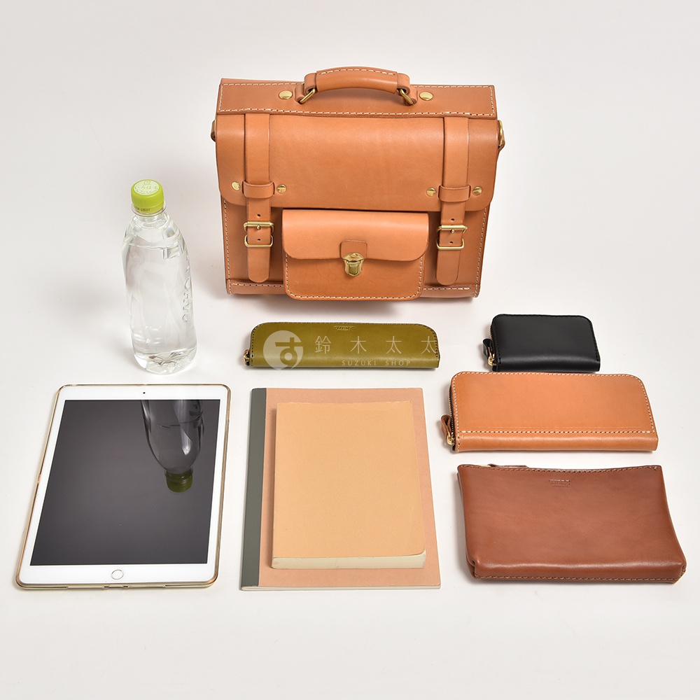 職人手作皮革學院風三用迷你後背包，以天然皮革製成，呈現經典棕色。包款設計有頂部提把、翻蓋式前袋，並以金屬釦環和皮帶固定。周圍擺放著平板電腦、筆記本、長夾、零錢包與綠色筆袋，展示其收納功能。