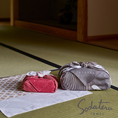 兩個以和風圖案風呂敷包裹的禮物，一個紅色，一個灰色，放在榻榻米上，旁邊有 Sodateru TOWEL 的品牌標誌。
