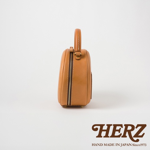 HERZ 品牌標誌，下方有 HAND MADE IN JAPAN Since1973 字樣，主體為一個棕褐色的皮革圓形手提包，從側面拍攝。