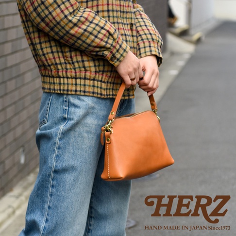 一位穿著格紋襯衫與牛仔褲的女性，手持一個由日本品牌 HERZ 職人手作的淺棕色皮革休閒包，包款設計簡約並配有金屬扣環與可拆式背帶，展現細膩的手工質感。