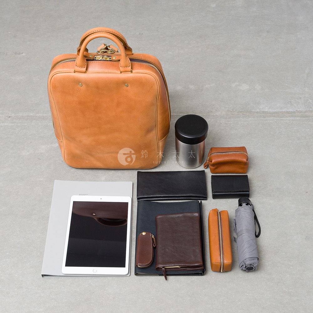 一個橘棕色的皮革後背包，搭配多個皮製小物，包括皮夾、筆袋、鑰匙包，以及一個平板電腦、一個不鏽鋼保溫杯和一把摺疊傘。