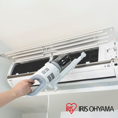 手持白色和灰色輕量吸塵器，正在清潔打開的白色空調內部。吸塵器具有灰色集塵筒和白色主機身，並有電線連接。