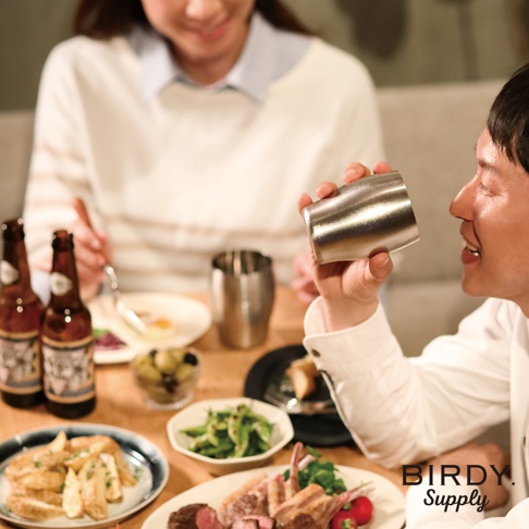 銀色不鏽鋼啤酒氣泡杯，握在男性手中，杯身帶有細緻的紋路，背景為餐桌上的食物、啤酒瓶和另一位女性。