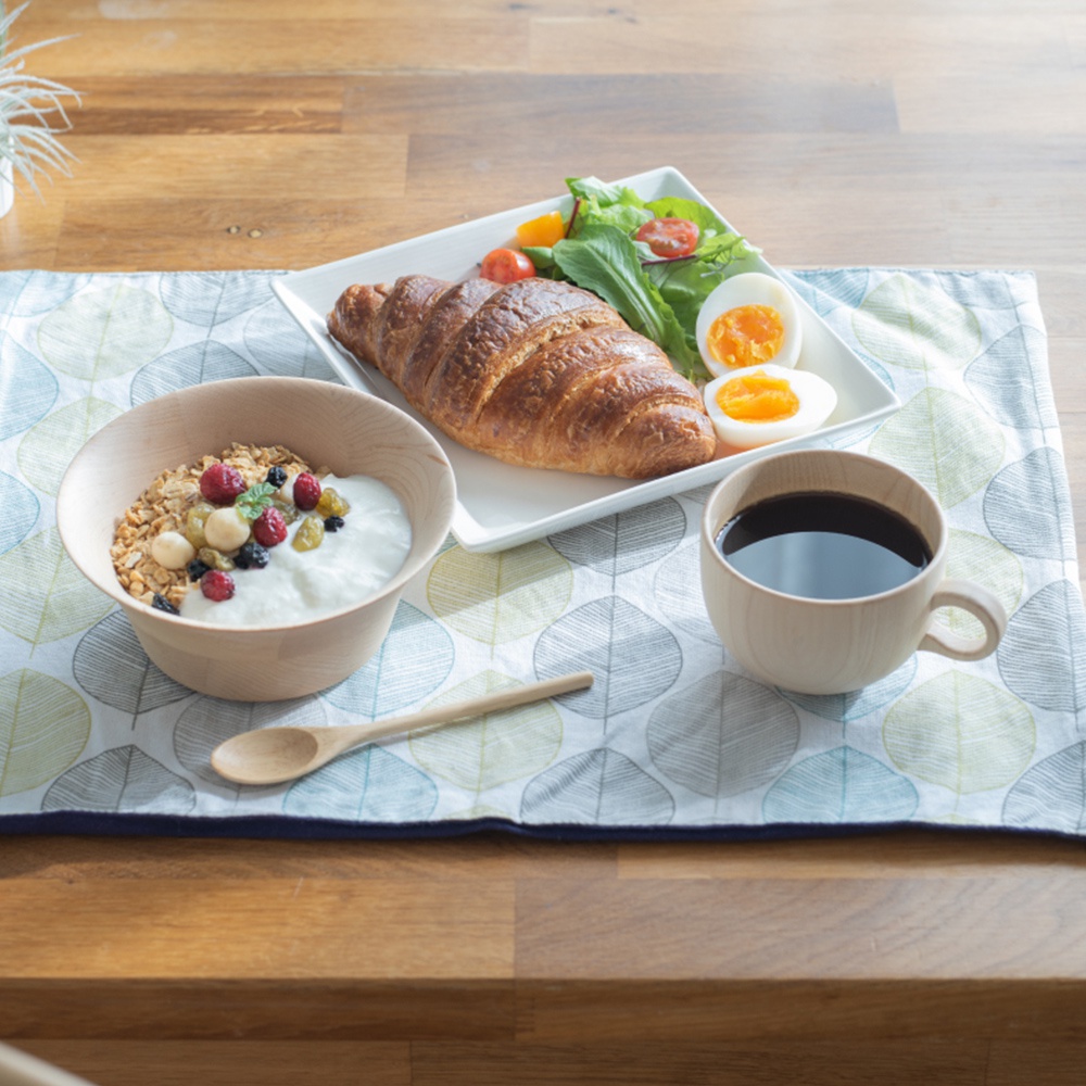 淺色楓木製咖啡杯，裝有深色咖啡，置於印有樹葉圖案的布墊上，旁邊是燕麥優格碗、可頌麵包與水煮蛋。