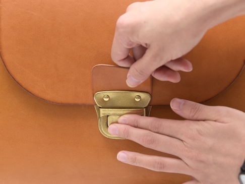 兩隻手正在操作一個棕褐色皮革包的黃銅鎖扣。皮革包表面光滑，帶有明顯的縫線。