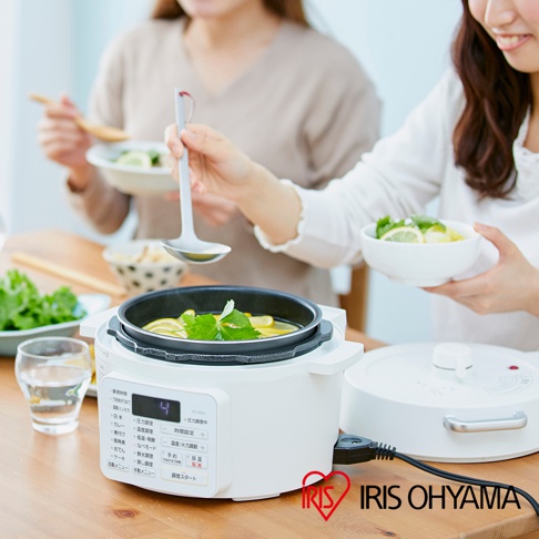 白色電子壓力鍋，內有檸檬與香草燉煮的湯品，鍋蓋旁有另一個白色鍋蓋，整體擺放在木質桌面，兩位女性在後方用餐。