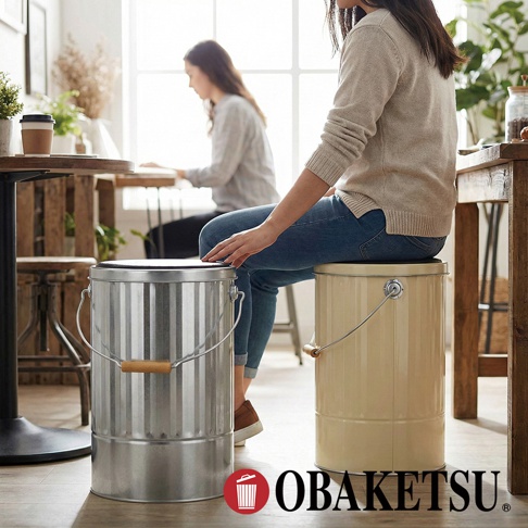 OBAKETSU 手工製凳子儲物桶 L 尺寸，一個為銀色鍍鋅鋼，另一個為奶油色，展示了其雙重用途，既是椅子也是收納桶。