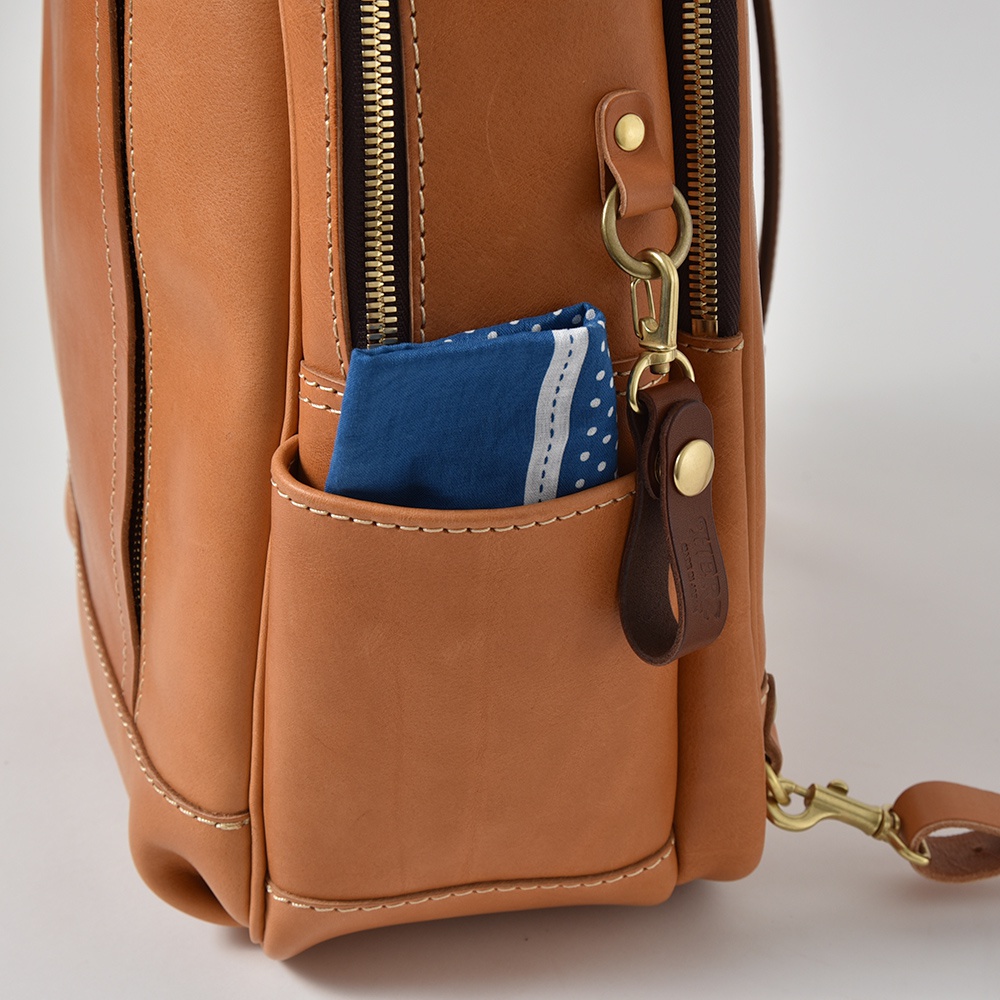 棕色皮革後背包，側面有一個外插袋，內有藍色印花方巾，並掛有黃銅色金屬扣環和深棕色皮革吊飾。
