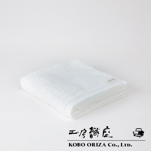 一張折疊的白色超細纖維浴巾，表面柔軟蓬鬆，邊緣車縫整齊，左上方角落有一個小小的品牌標籤，背景為純白色。