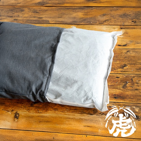 灰色與白色布料拼接的長枕，一側為素面灰色，另一側為帶有細緻紋路的淺灰色或白色，枕頭置於木紋背景上。