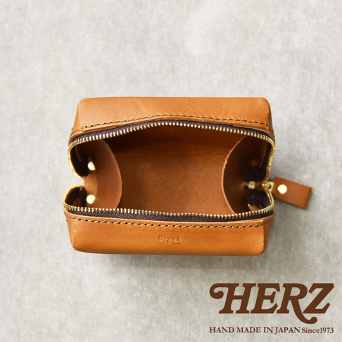 HERZ 品牌標誌，下方有 HAND MADE IN JAPAN Since1973 字樣，上方為一個打開的棕色皮革箱型收納包，內部空間可見，拉鍊為金色，包包表面壓印有 Organ 字樣。